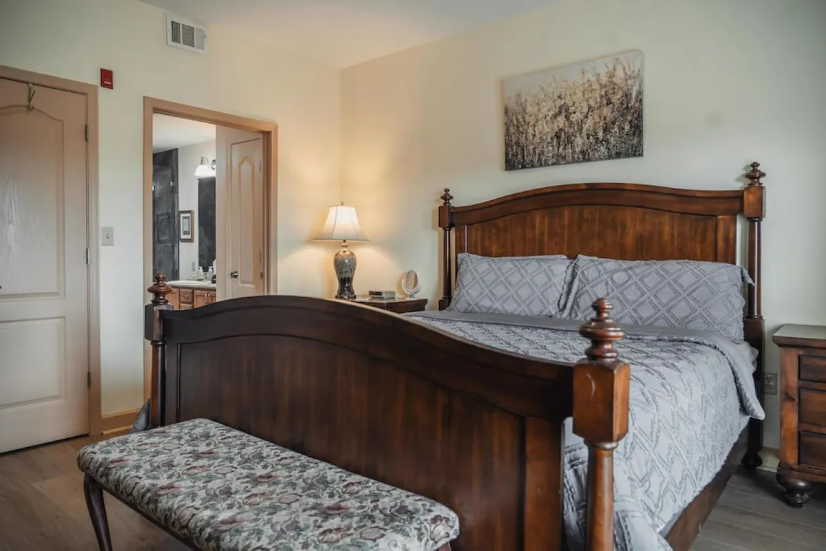 The image shows a serene and inviting bedroom featuring a wooden bed with plush linens, a decorative bench at the foot, a nightstand with a softly glowing lamp, and calming mountain-inspired artwork on the wall. Natural light filters gently into the space, creating a cozy and restful atmosphere ideal for unwinding after a day of adventure.