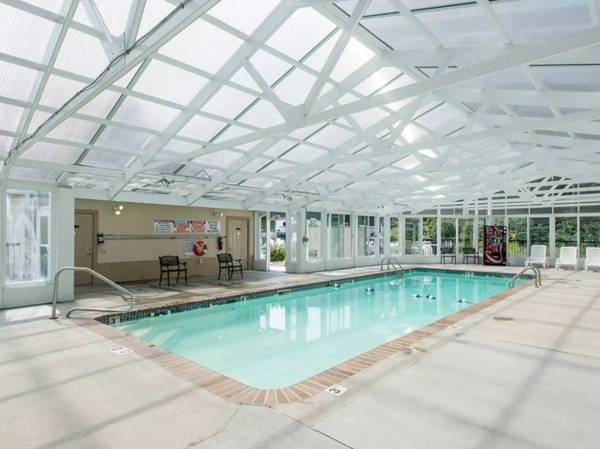A tranquil interior view of a spacious indoor pool, basked in natural light from large windows, with comfortable lounge chairs lining the poolside tiles. The white beams and clean design evoke a year-round oasis for swimming and relaxation.