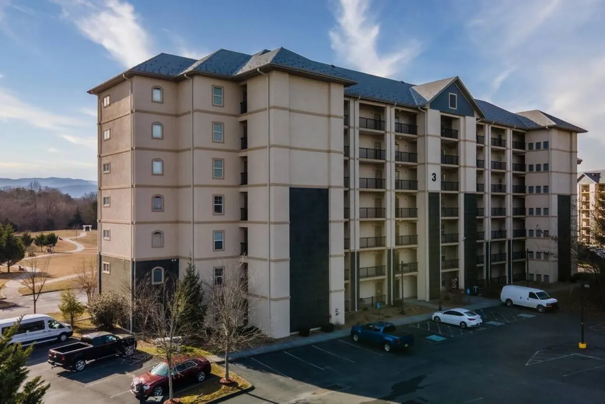 The exterior of a multi-story condo building framed by a lush landscape and spacious parking lot. The scene looks inviting and accessible, with plenty of parking available for guests.