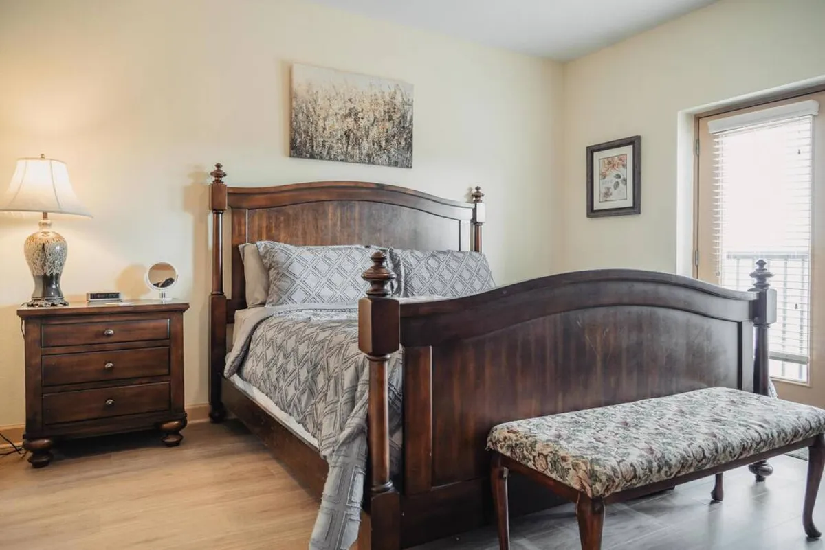 A peaceful master bedroom featuring a wooden bed frame, a decorative bench at the foot of the bed, and a nightstand with a lamp. Natural light streams in, creating a relaxing retreat ideal for rest and privacy.
