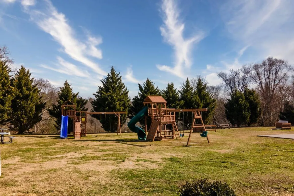 A vibrant playground scene with colorful swings, a slide, and green grass set among trees. The area feels safe, spacious, and inviting for children, with ample room for parents to relax or supervise nearby.