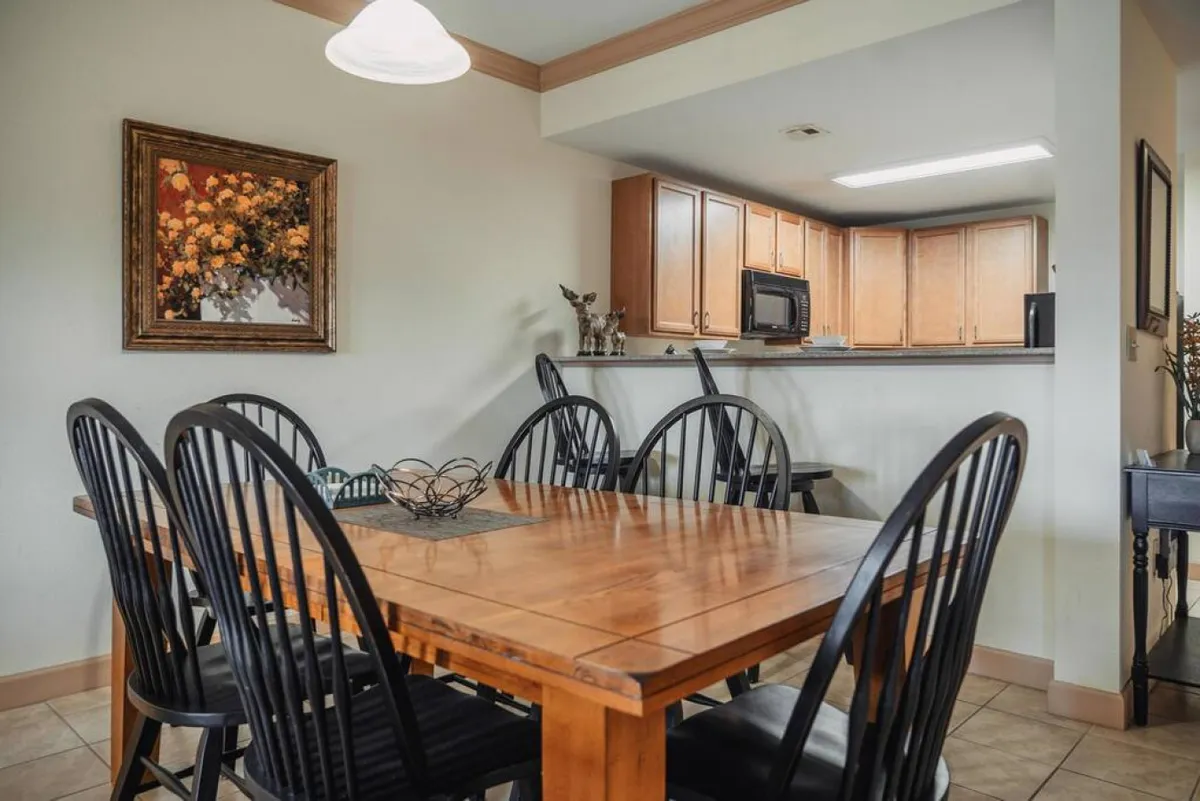 A spacious, light-filled dining area with a wooden table, black chairs, and a view into the kitchen area. Framed artwork and warm lighting create an inviting atmosphere ideal for group meals or game nights.