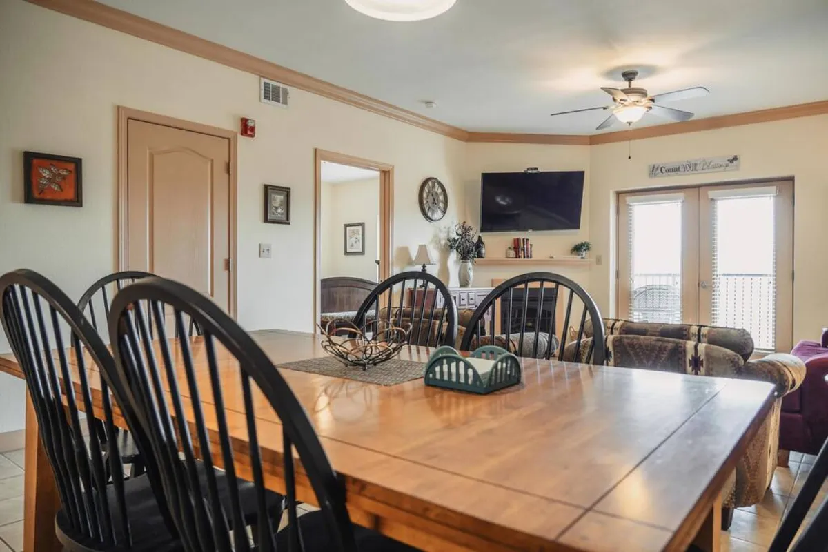 An open-concept dining and living area with a central dining table, comfortable seating, a flat-screen TV, and doors leading outside. The setting invites family meals, TV nights, and relaxing gatherings.