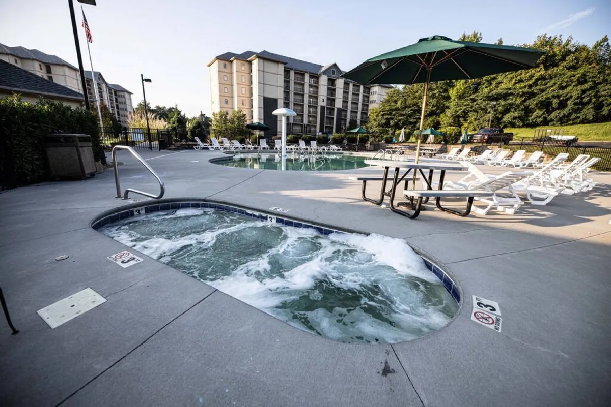 An inviting exterior shot featuring a sparkling swimming pool, relaxing hot tub, lounge chairs, picnic table, and umbrella, all arranged in a sunny, resort-like setting. The scene suggests relaxation, socializing, and outdoor enjoyment for families or groups.