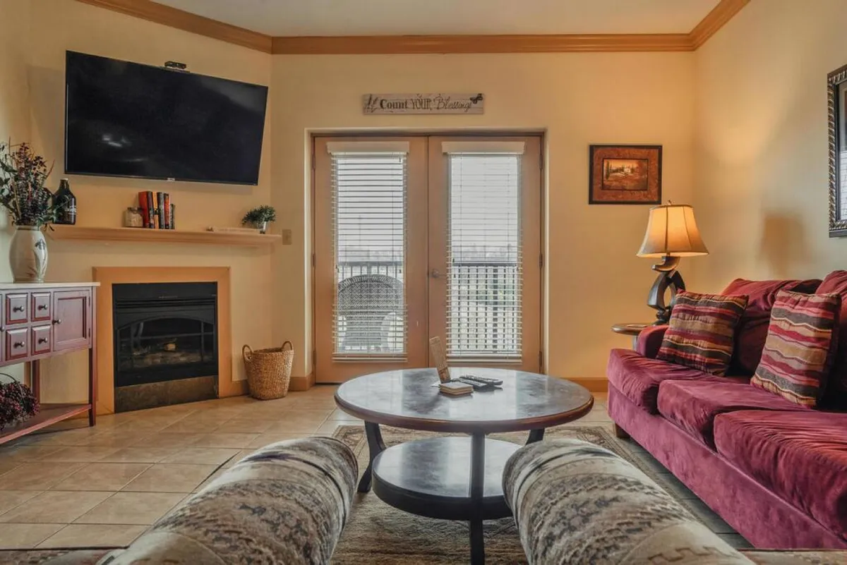A welcoming living room with a fireplace, comfortable sofa, coffee table, TV, and a door leading to the balcony. The space is arranged for relaxation, socializing, or family movie nights.